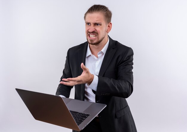 Handsome business man wearing suit holding laptop computer looking frustrated with angry face standing over white background