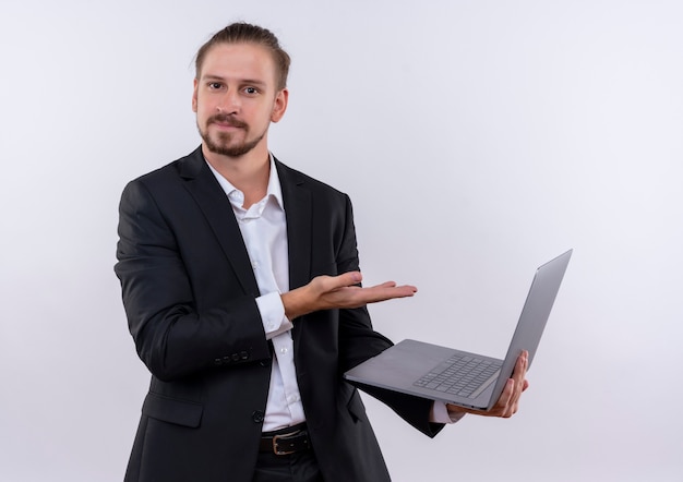 Handsome business man wearing suit holding laptop computer presenting with arm of hand looking confident standing over white background