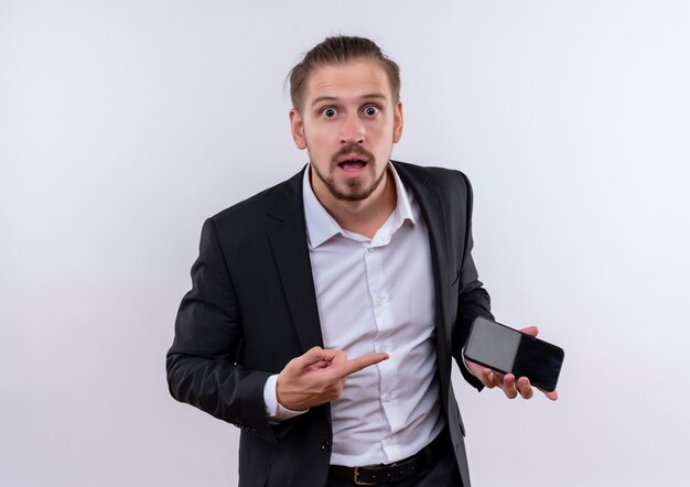 Handsome business man wearing suit holding smartphone pointing with finger to it looking surprised and amazed standing over white background