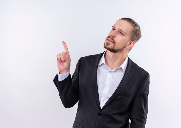 Handsome business man wearing suit looking aside with pensive expression pointing with finger up standing over white background