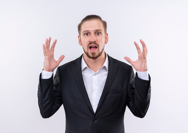 Free photo handsome business man wearing suit looking at camera disappointed raising arms standing over white background