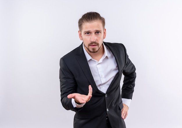 Handsome business man wearing suit looking at camera with arm out as asking question standing over white background