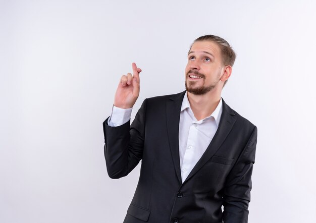 Handsome business man wearing suit making desirable wish crossing fingers smiling standing over white background