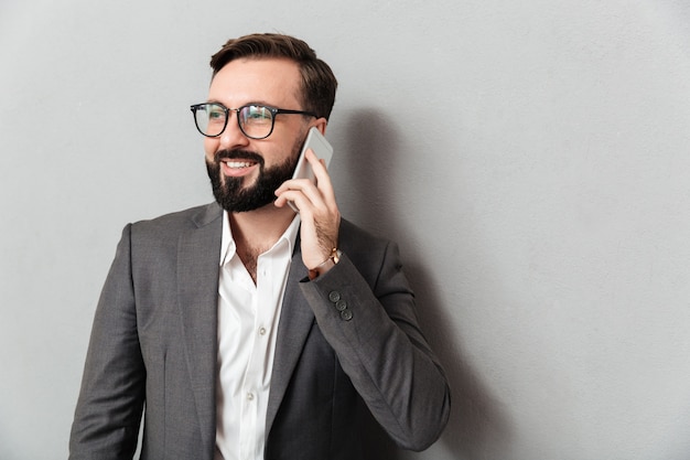 Handsome businesslike man in formal wear having mobile conversation using smartphone isolated over gray