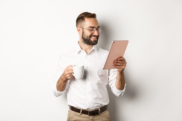 Handsome businessman drinking coffee and reading on digital tablet, smiling pleased, standing  