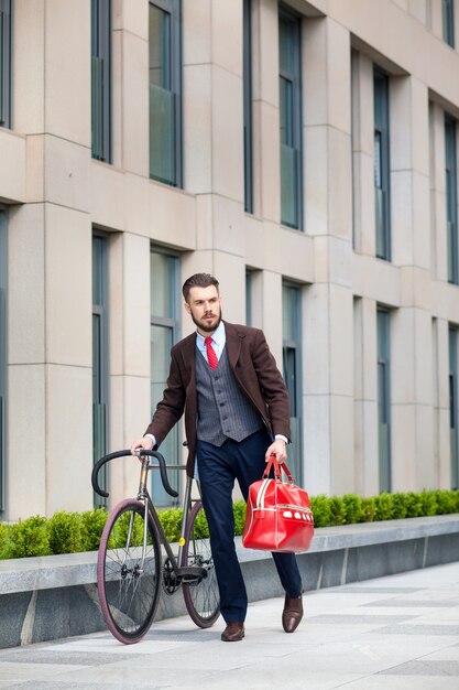 Handsome businessman in a jacket and red bag and his bicycle on city streets. 