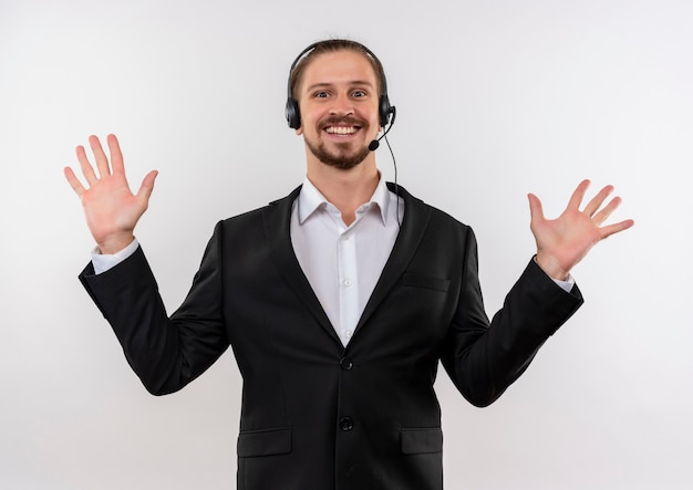 Handsome businessman in suit and headphones with a microphone looking at camera raising palms in surrender standing over white background