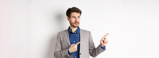 Free photo handsome businessman in suit pointing looking left at logo with serious face standing against white