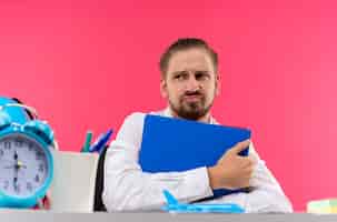 Free photo handsome businessman in white shirt holding folder looking aside with sad expression sitting at the table in offise over pink background