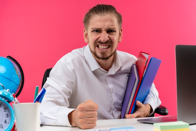 Handsome businessman in white shirt holding folders looking at camera with angry face sitting at the table in offise over pink background