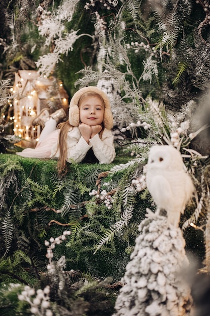 Handsome caucasian child with long fair hair lies in christmas atmosphere with a lot of decorate trees around her and owl