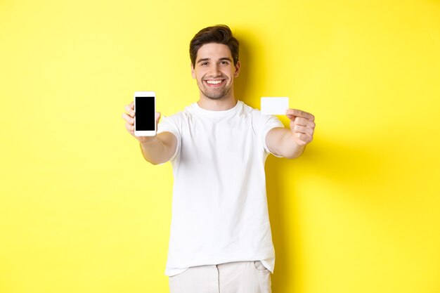 Handsome caucasian male model showing smartphone screen and credit card, concept of mobile banking and online shopping, yellow background.