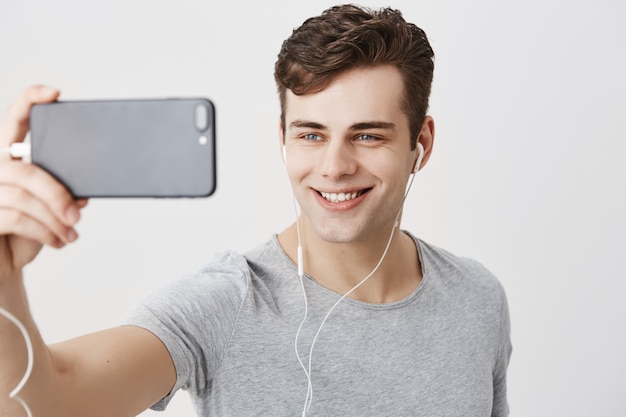 Free photo handsome caucasian young male with dark hair and appealing blue eyes holding mobile phone, posing for selfie, looking  with flirty smile. good-looking guy making video call.