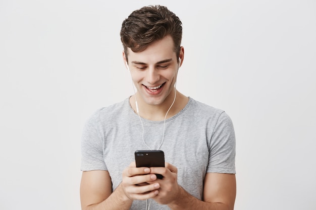 Free Photo handsome caucasian young male with dark hair and blue eyes holding mobile phone, smiling with teeth, typing message to his girlfriend, wearing earphones. people, lifestyle and modern communication.