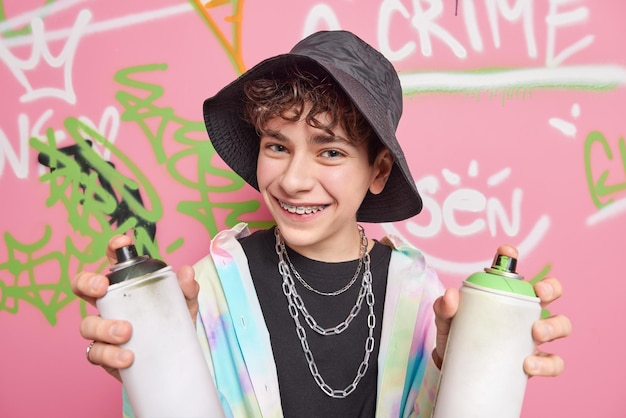 Free photo handsome cheerful hipster boy poses in city against graffiti background wears black panama shirt silver chains braces on teeth holds two cans of paint enjoys favorite hobby street art concept