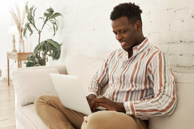 Handsome cheerful young dark skinned male smm manager enjoying working from home sitting on sofa with laptop computer