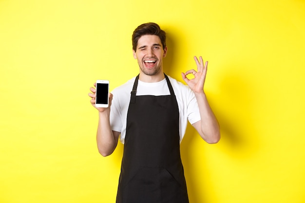 Handsome coffee shop worker showing ok sign and smartphone screen, recommending application, standing over yellow background