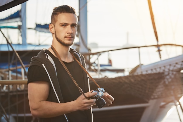 Handsome confident guy with stylish haircut standing near awesome yacht, holding camera, staring seriously and being focused during photo session in harbour, making pictures of scenery