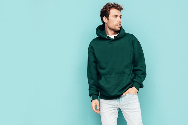 Handsome confident hipster modelSexy unshaven man dressed in summer stylish green hoodie and jeans clothes Fashion male with curly hairstyle posing in studio Isolated on blue