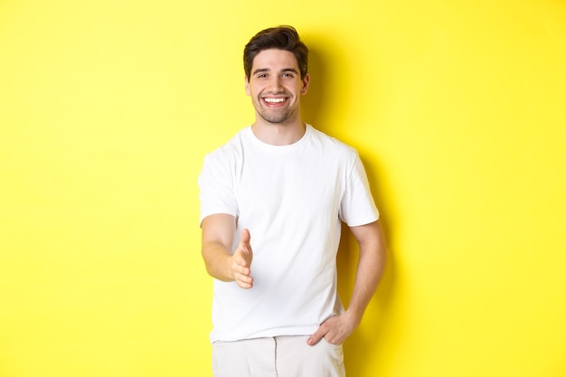 Handsome and confident man extending hand for handshake, greeting you, saying hello, standing in white t-shirt over yellow background.