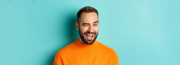 Free Photo handsome confident man winking at camera smiling sassy standing in orange sweater against blue backg