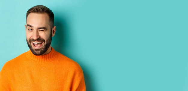 Free photo handsome confident man winking at camera smiling sassy standing in orange sweater against blue backg