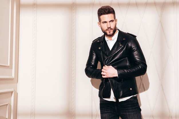 Handsome confident model Sexy stylish man in biker leather jacket and black jeans Fashion hipster male posing near white wall in studio interior at sunny day Shadow from window