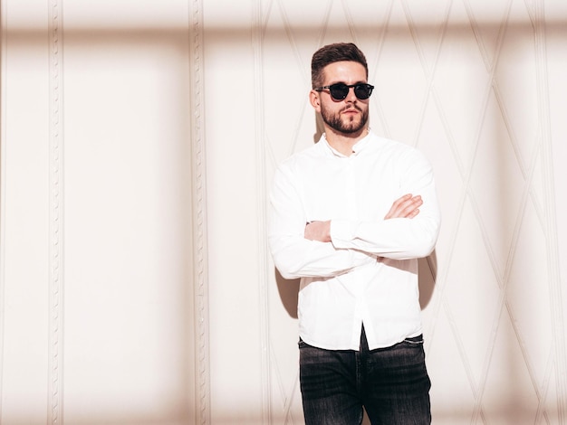 Handsome confident model Sexy stylish man dressed in white shirt and jeans Fashion hipster male posing near white wall in studio interior at sunny day Shadow from window In sunglasses