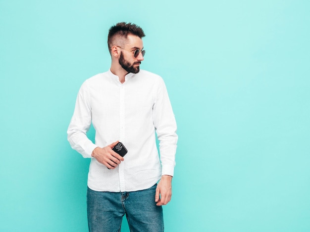 Free Photo handsome confident modelsexy stylish man dressed in shirt and jeans fashion hipster male posing near blue wall in studio holding smartphone looking at cellphone screen using apps isolated