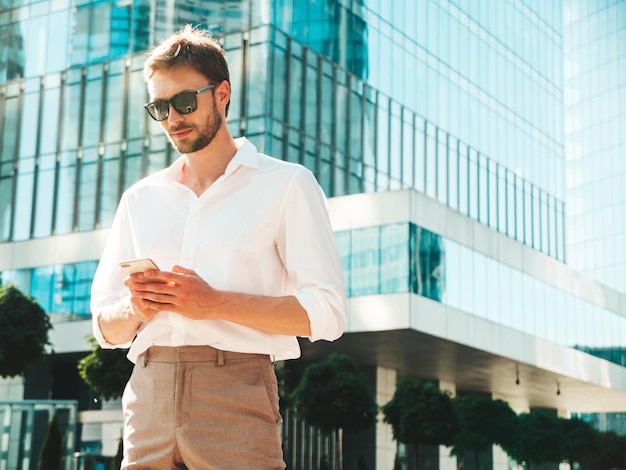 Handsome confident stylish hipster lambersexual modelModern man dressed in white shirt Fashion male posing on the street background near skyscrapers Outdoors at sunset Using smartphone apps