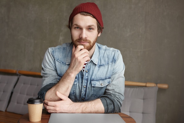 Free photo handsome confident young caucasian bearded freelancer in jeans jacket and hat