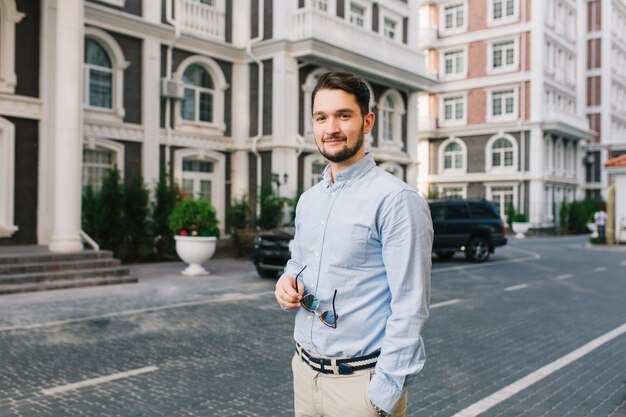 Handsome dark-haired man in blue shirt is walking around British quarter. He holds sunglasses