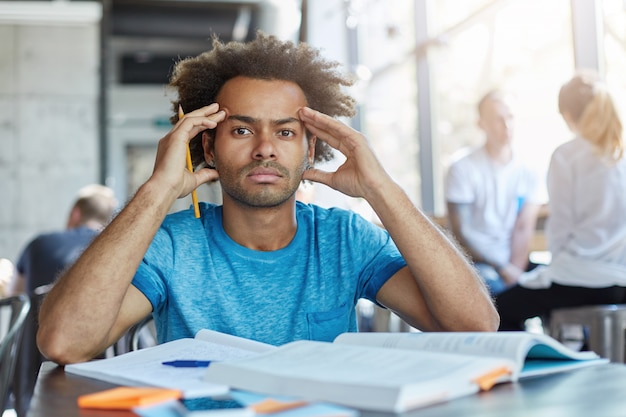 Handsome dark-skinned bearded student with Afro haircut squeezing temples, having headache while preparing for exams or tests night and day, looking with painful frustrated expression