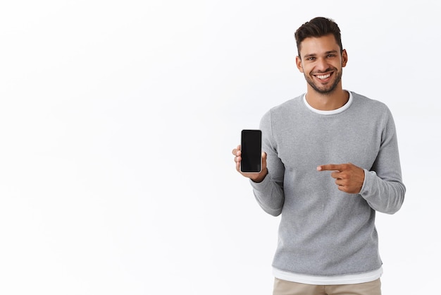 Free photo handsome enthusiastic smiling man with bristle in grey sweater holding smartphone screen facing camera pointing mobile display and grinning recommend application carsharing or shopping site