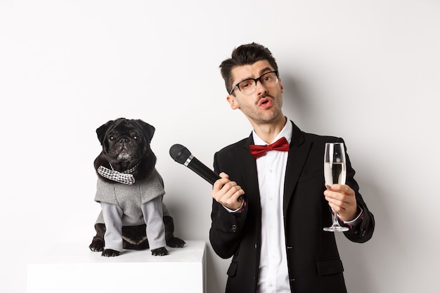 Handsome fancy man in glasses, raising glass of champagne and giving microphone to cute pug in party suit, celebrating and having fun, white background