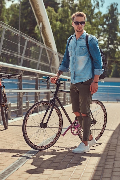 Free photo handsome fashionable hipster with a stylish haircut in sunglasses walking with bicycle outdoors.