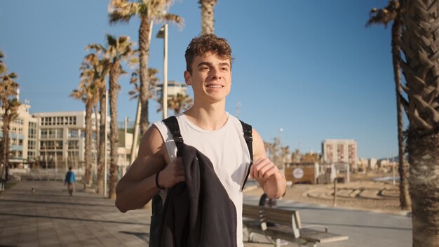 Handsome fit guy with backpack happily walking along seaside after workout outdoor