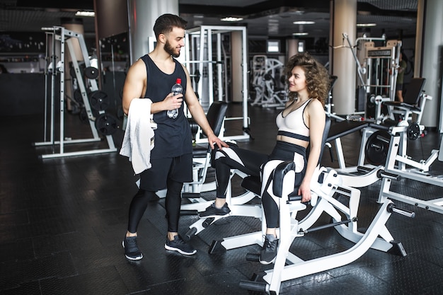 Handsome fitness instructor is helping his attractive client how to work out on an exercise in gym