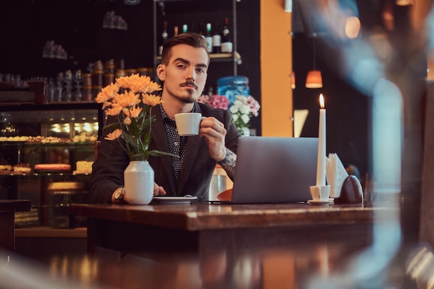 Handsome freelancer man with stylish beard and hair dressed in a black suit sitting at a cafe with an open laptop and holds a cup of coffee, looking at a camera.