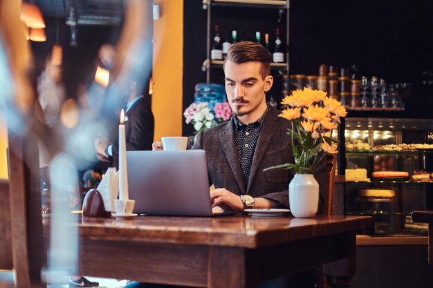 Handsome freelancer man with stylish beard and hair dressed in a black suit working on a laptop while sitting at a cafe.