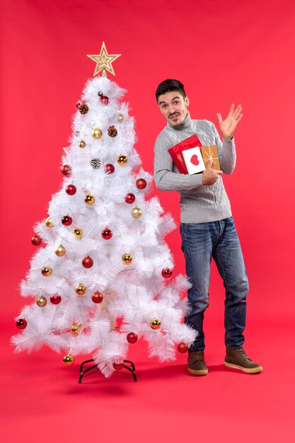 Handsome funny young man standing near the decorated white New Year tree and holding his gifts