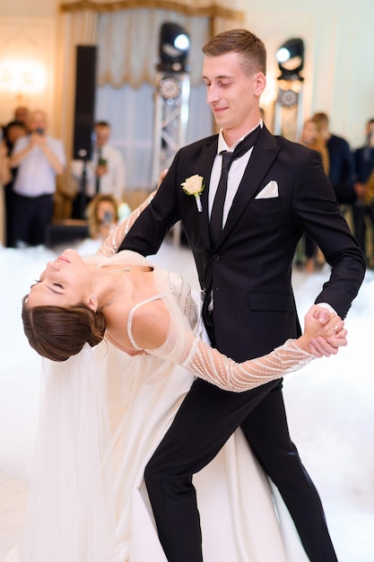 Free photo handsome groom in black stylish suit holding hands of bride woman in beautiful wedding dress while dancing in the restaurant attractive bride girl in long sleeves dress loving married couple