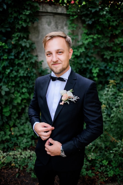 Free photo handsome groom in black suit and white rose boutonniere stands in the garden