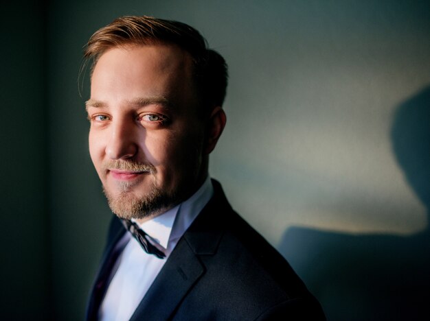 Handsome groom in classy black suit stands in a dark room