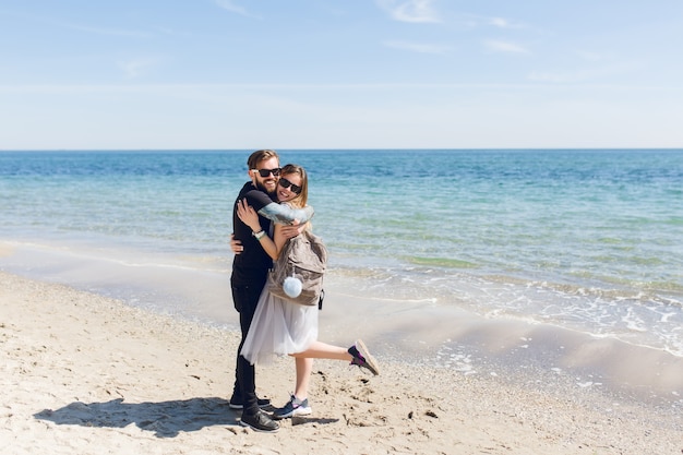 Handsome guy in black T-shirt and pants is hugging pretty woman with long hair near sea