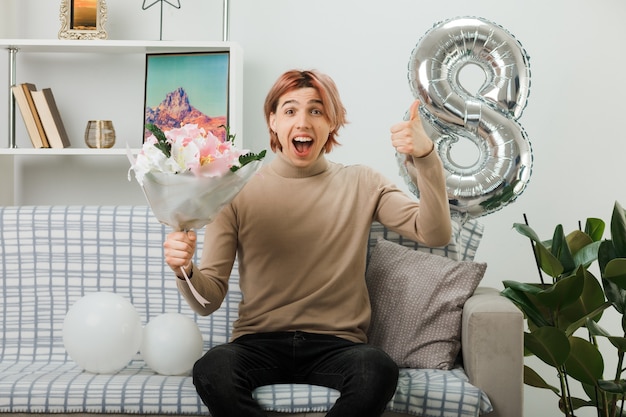 Handsome guy on happy women day holding bouquet sitting on sofa in living room