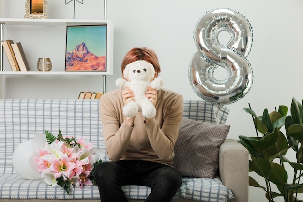 Handsome guy on happy women day holding and covered face with teddy bear sitting on sofa in living room