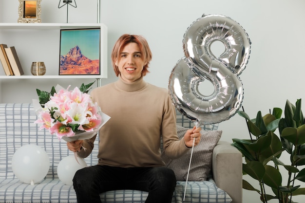 Handsome guy on happy women day holding number eight balloon and bouquet sitting on sofa in living room