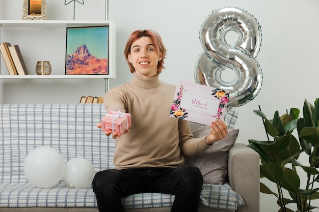 Handsome guy on happy women day holding out present with postcard at camera sitting on sofa in living room