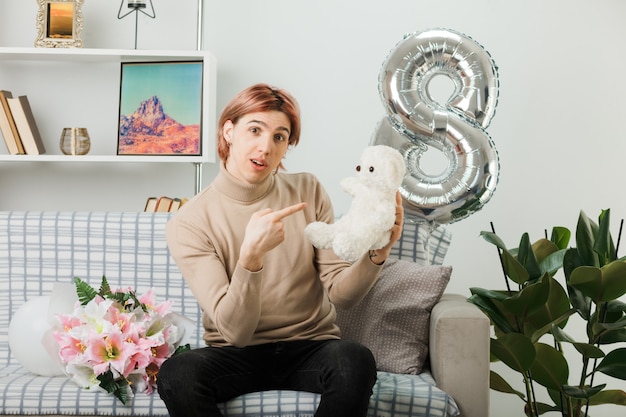 Handsome guy on happy women day holding and points at teddy bear sitting on sofa in living room